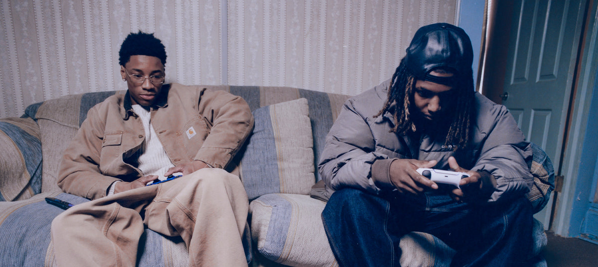 Two people sitting on a couch wearing Carhartt WIP clothing, both holding remote controls, in a room.