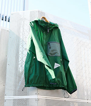 Vivid green And Wander jacket hanging against urban architecture, blending outdoor functionality with striking contemporary style.
