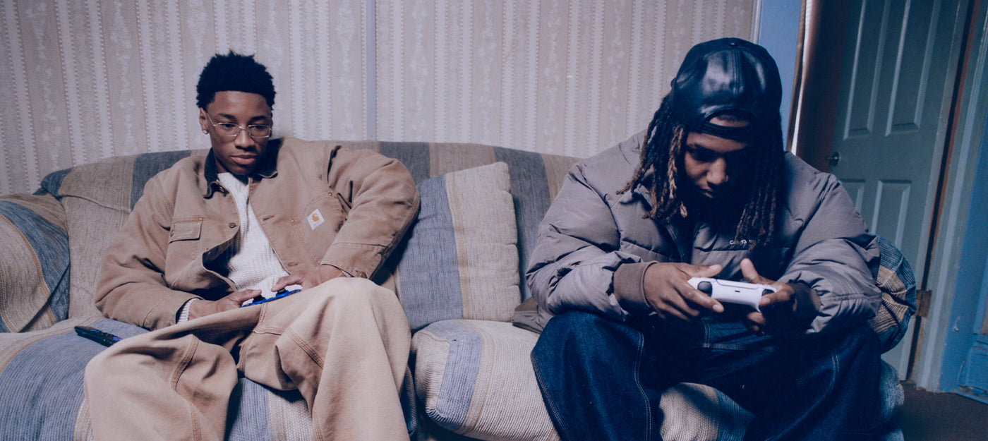 Two people sitting on a couch wearing Carhartt WIP clothing, both holding remote controls, in a room.